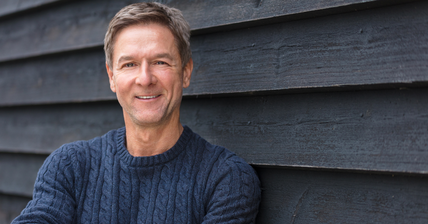 Smiling mature man in a navy cable-knit sweater leaning against a wooden wall