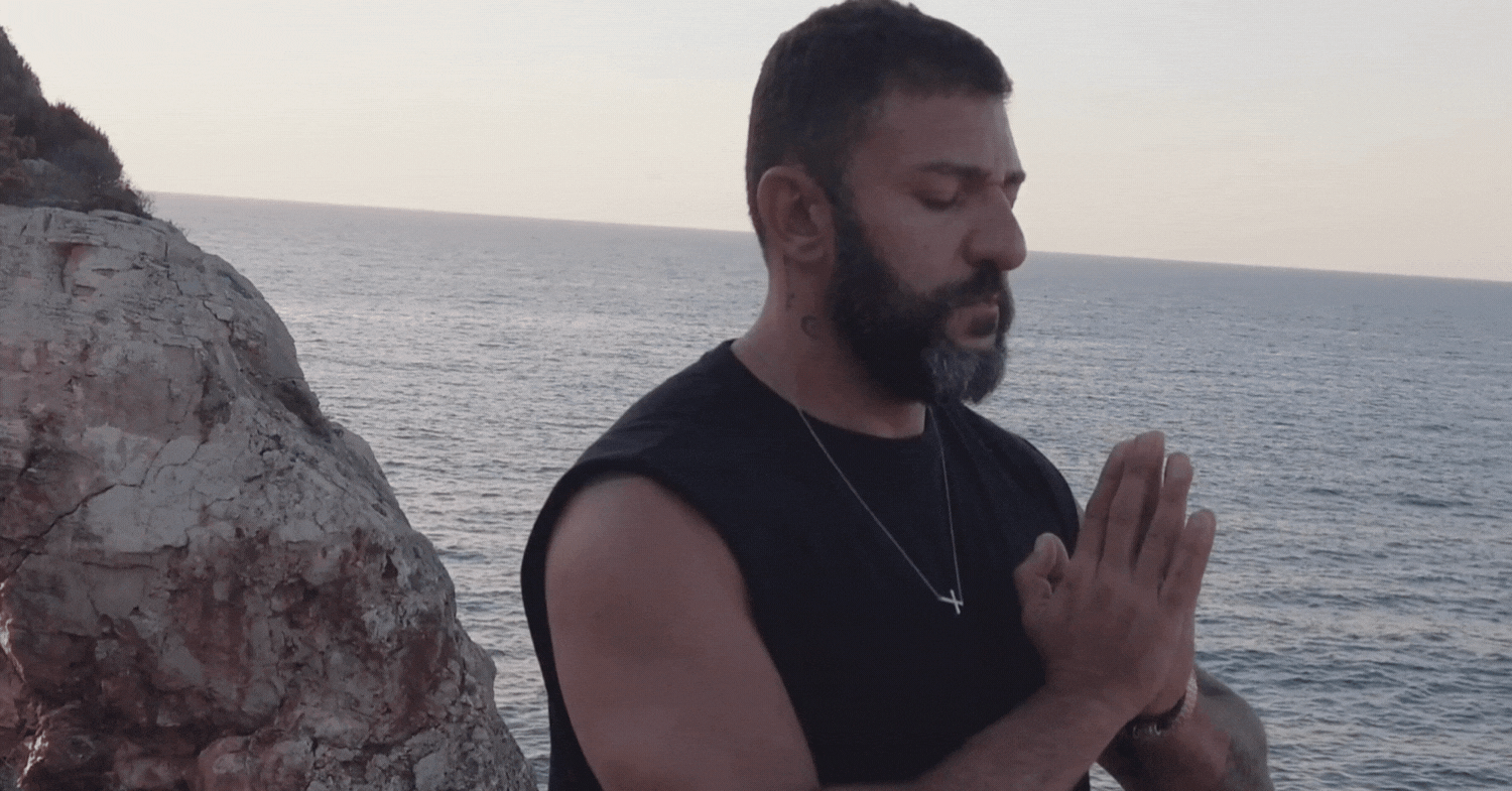 Bearded man meditating with his hands pressed together beside the sea