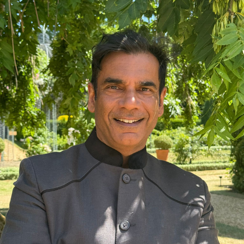 Dr Ash Kapoor smiling outdoors in a garden, wearing a grey collarless jacket
