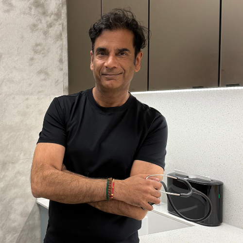 Dr Ash Kapoor standing with folded arms in a black T-shirt beside a clinic worktop, holding a stethoscope