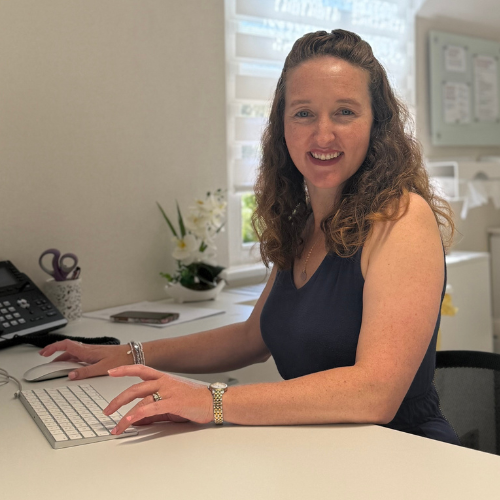 Dr Caitlin Scott smiling at a desk with a keyboard and telephone, wearing a navy sleeveless top