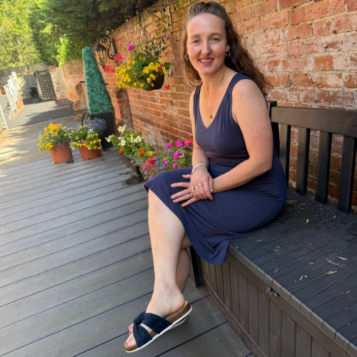 Dr Caitlin Scott sitting on a bench on a decked terrace beside a brick wall with hanging flower baskets