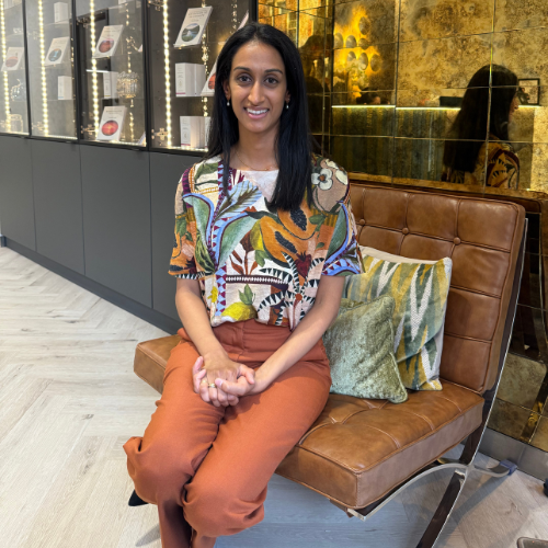 Dr Madhvi Patel sitting on a tan leather chair in a clinic lounge, wearing a patterned blouse and rust-coloured trousers