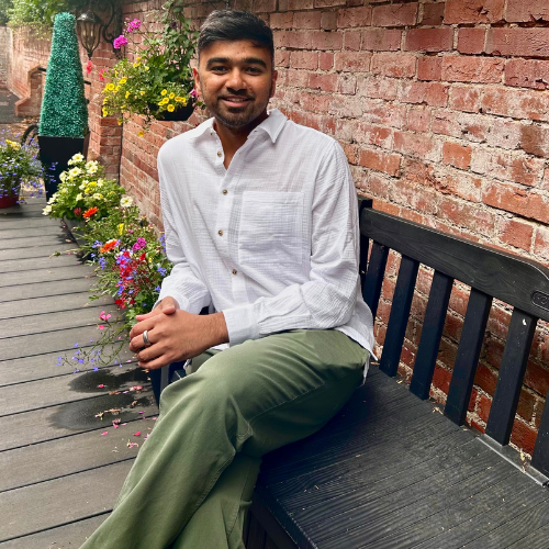 Dr Vijay Prakash sitting on a bench on a decked terrace beside a brick wall with hanging flower baskets, wearing a white shirt