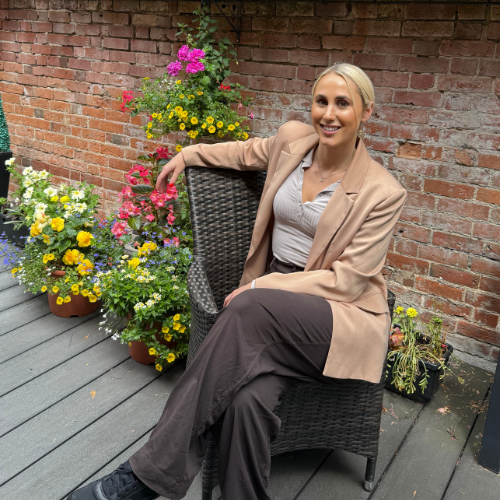 Cara Richardson sitting in a wicker chair on a decked terrace among potted flowers beside a brick wall