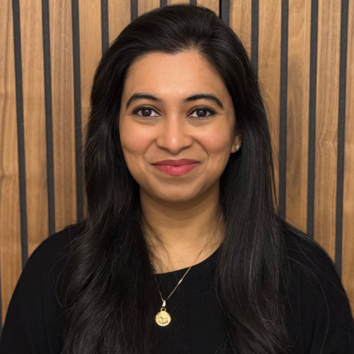 Dr Gunjan Modi smiling, with long dark hair, wearing a black top and a gold pendant, in front of wooden slats