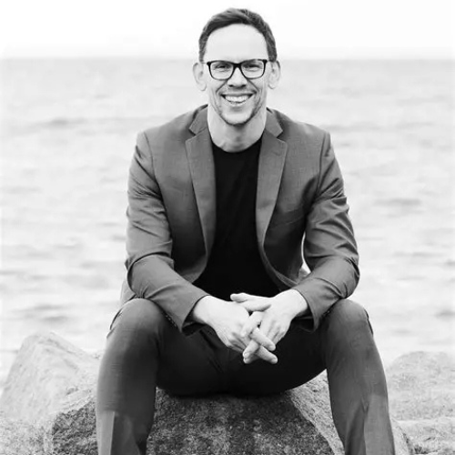Black and white photograph of Dr Steve James sitting on a rock by the sea, wearing glasses and a blazer