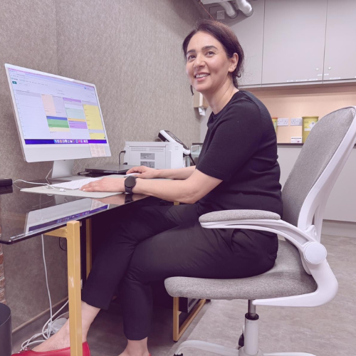 Dr Kiran Dhillon smiling at a desk with a computer in a clinic consulting room