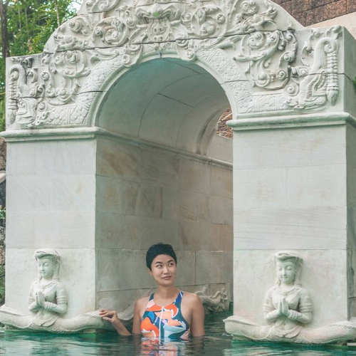 A woman in a swimming costume in a pool beneath a carved white stone arch flanked by statues