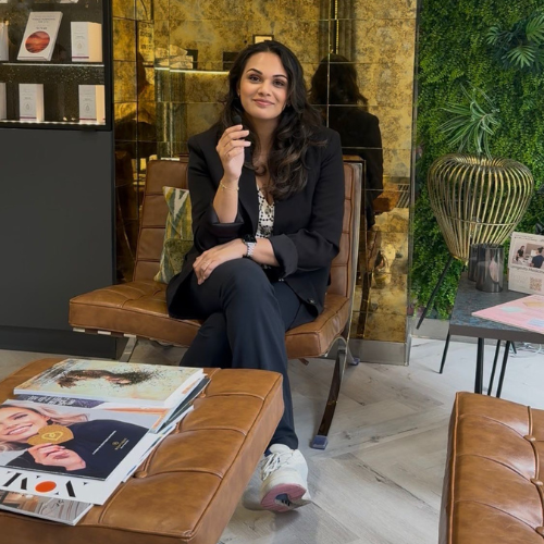 Nikita Desai sitting on a tan leather chair in a clinic lounge, wearing a black trouser suit