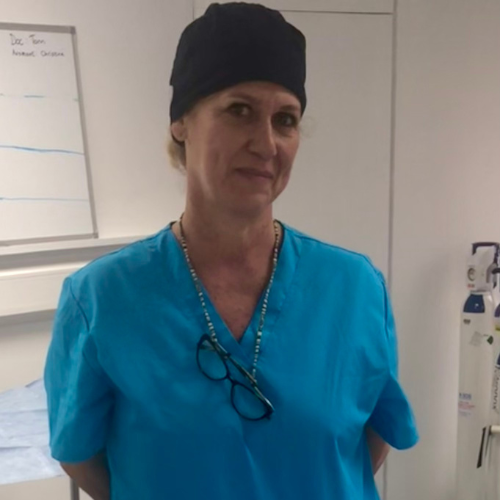 Christina Walker in turquoise scrubs and a black scrub cap, glasses hanging on a cord around her neck, in a clinic room