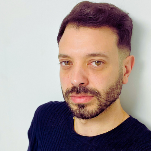 Dr Moe Irshidat, with a beard, wearing a navy jumper against a pale wall, his hair tinted purple