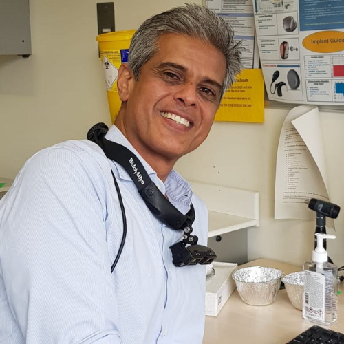 Hamid Daya smiling in a clinic room, a headlight on a strap around his neck, wearing a light blue shirt