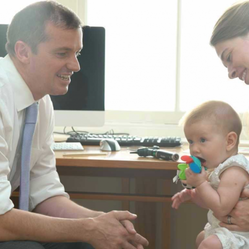 Ian Lilly smiling at a baby held on a woman’s lap beside his consulting room desk